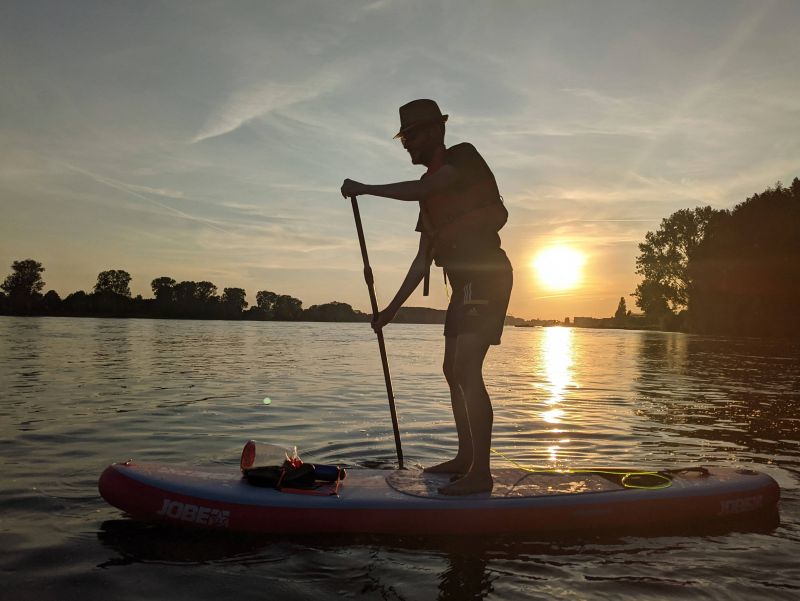 Ein Stehpaddler auf dem Rhein bei Bonn. Es sind die Silhouetten der Person und von Bäumen erkennbar, im Hintergrund geht die Sonne unter. Bild: privat