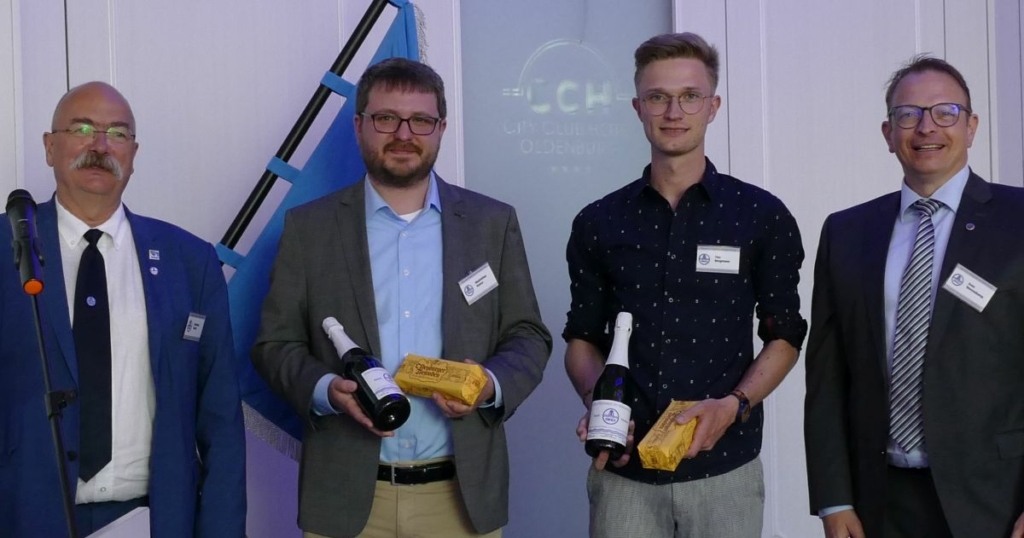 Vier Männer stehen nebeneinander. Die beiden in der Mitte halten jeweils eine Flasche Sekt und eine gelbe Packung in der Hand.