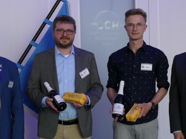 Vier Männer stehen nebeneinander. Die beiden in der Mitte halten jeweils eine Flasche Sekt und eine gelbe Packung in der Hand.